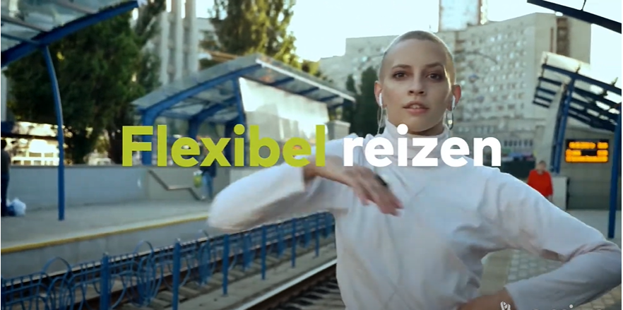 Vrouw met gemillimeterd haar in witte coltrui maakt een dansbeweging en kijkt in de camera terwijl ze op een perron staat. Met de tekst 'Flexibel reizen'.