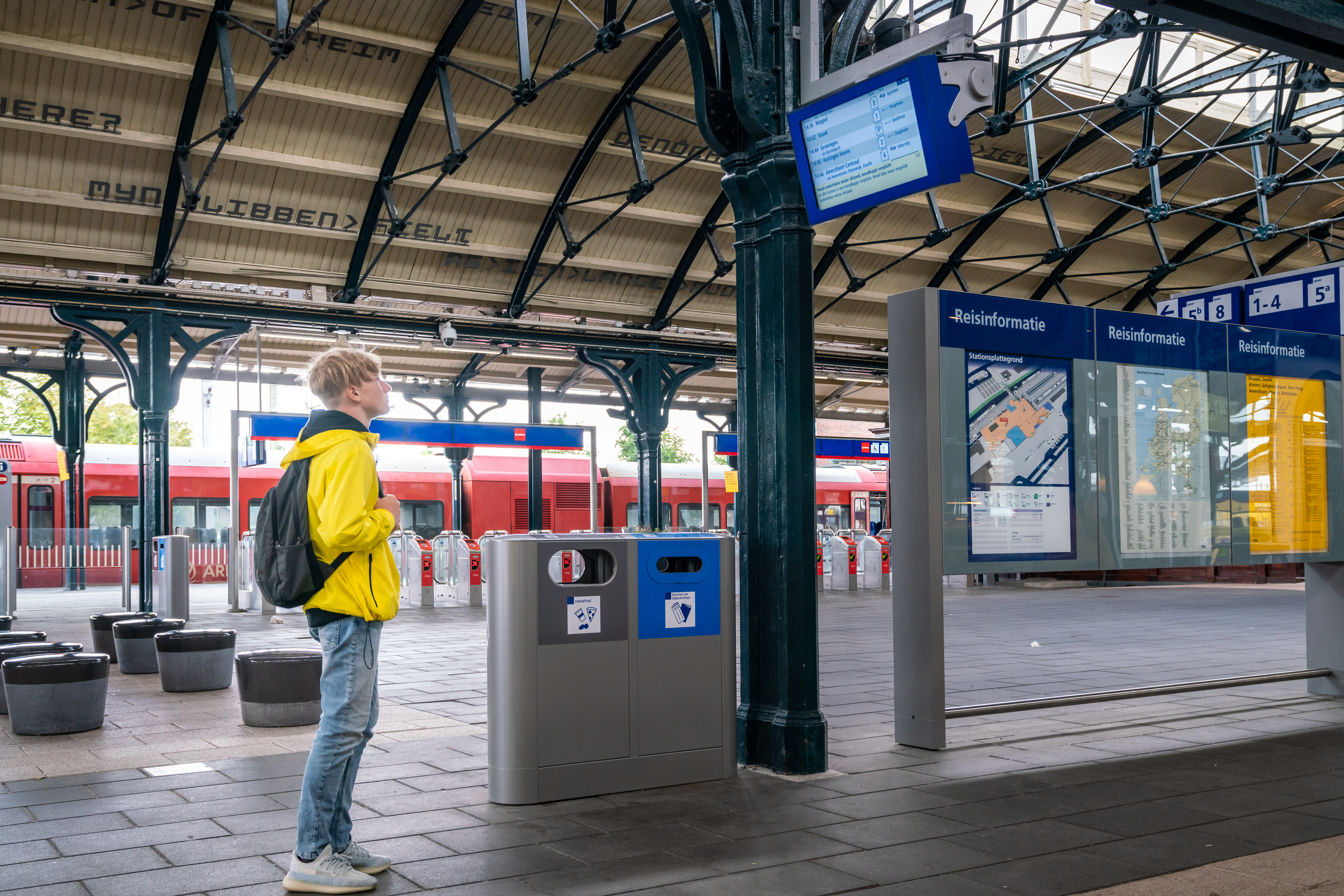 Tiener in felgele jas staat naar de vertrektijden te kijken op een treinperron. Op de achtergrond staat een rode Arriva-trein.