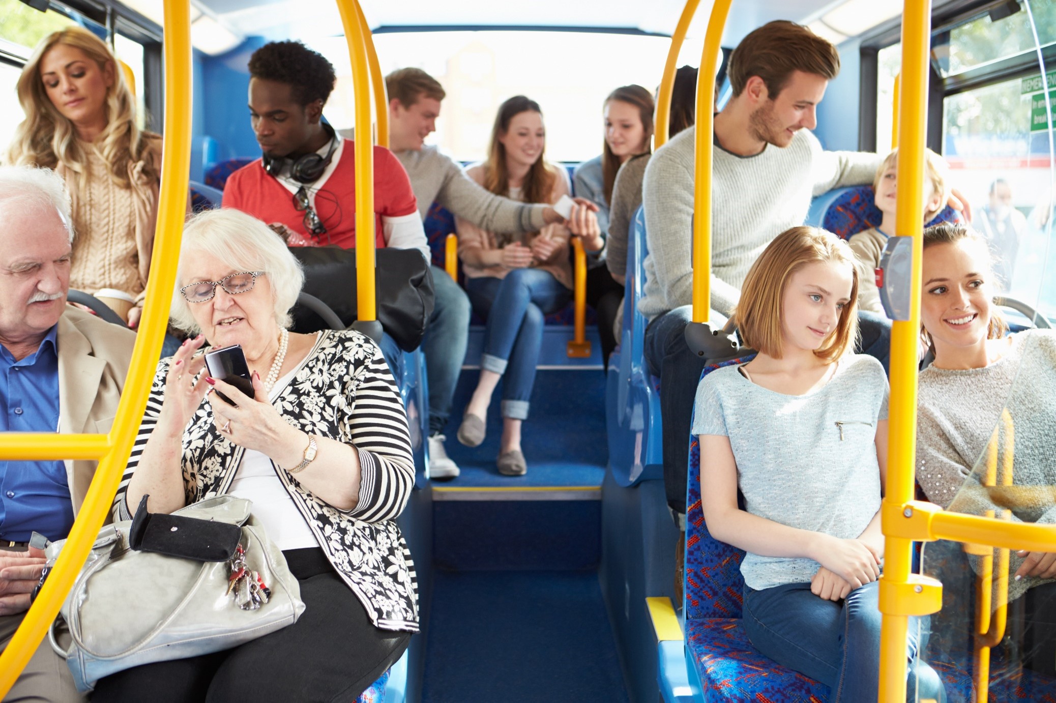 Achterste drie rijen stoelen in zonnige Arriva-bus, gevuld met diverse reizigers. Oud stel, man met klein kind, twee vriendinnen en een groepje vrienden creëren een positieve sfeer.