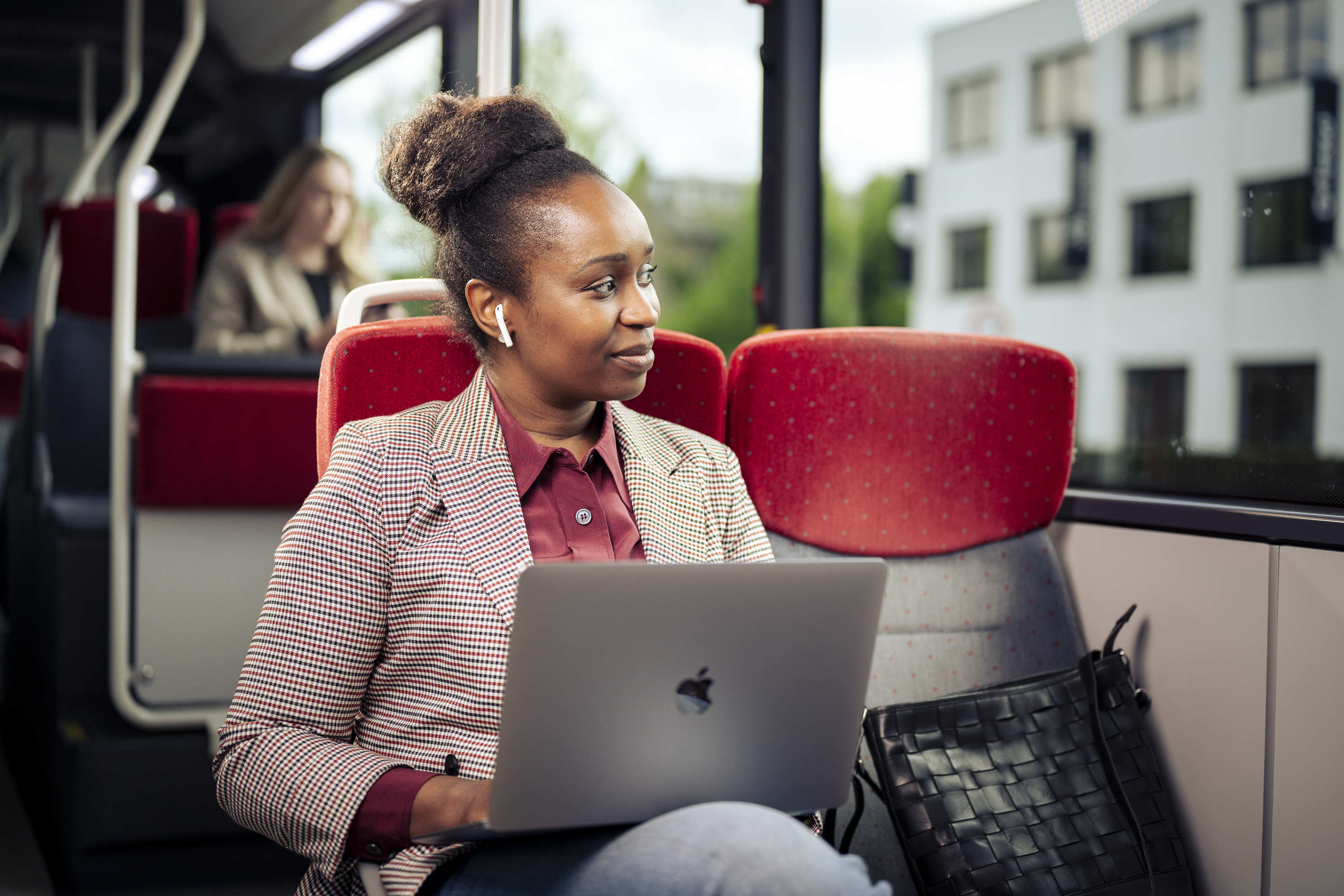 Een vrouw zit in de bus in pak met oordopjes in en laptop op schoot terwijl ze uit het raam kijkt.
