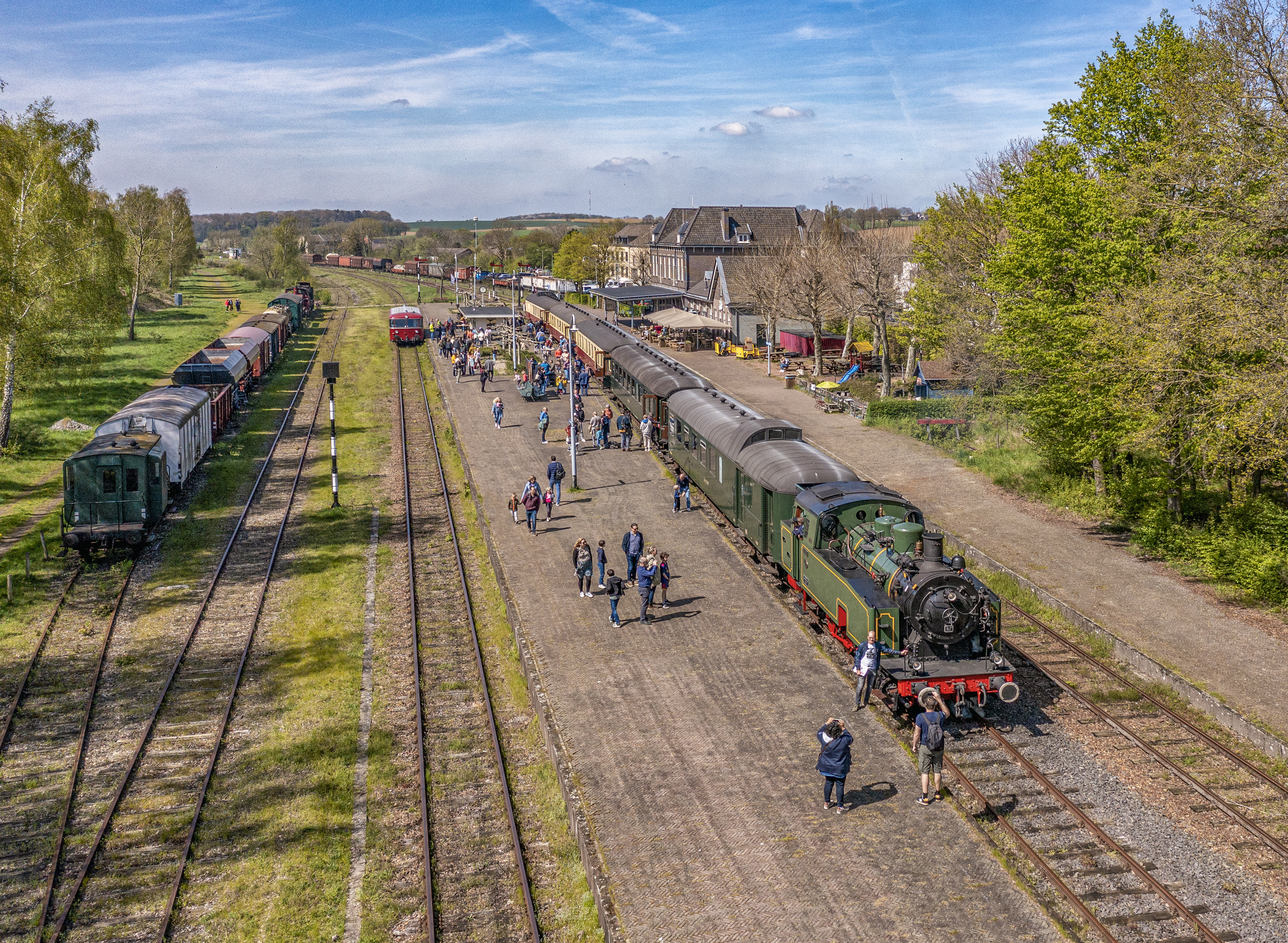 Een groen stoomtrein met zes wagons staat op station Simpelveld, er lopen allemaal mensen langs de trein.