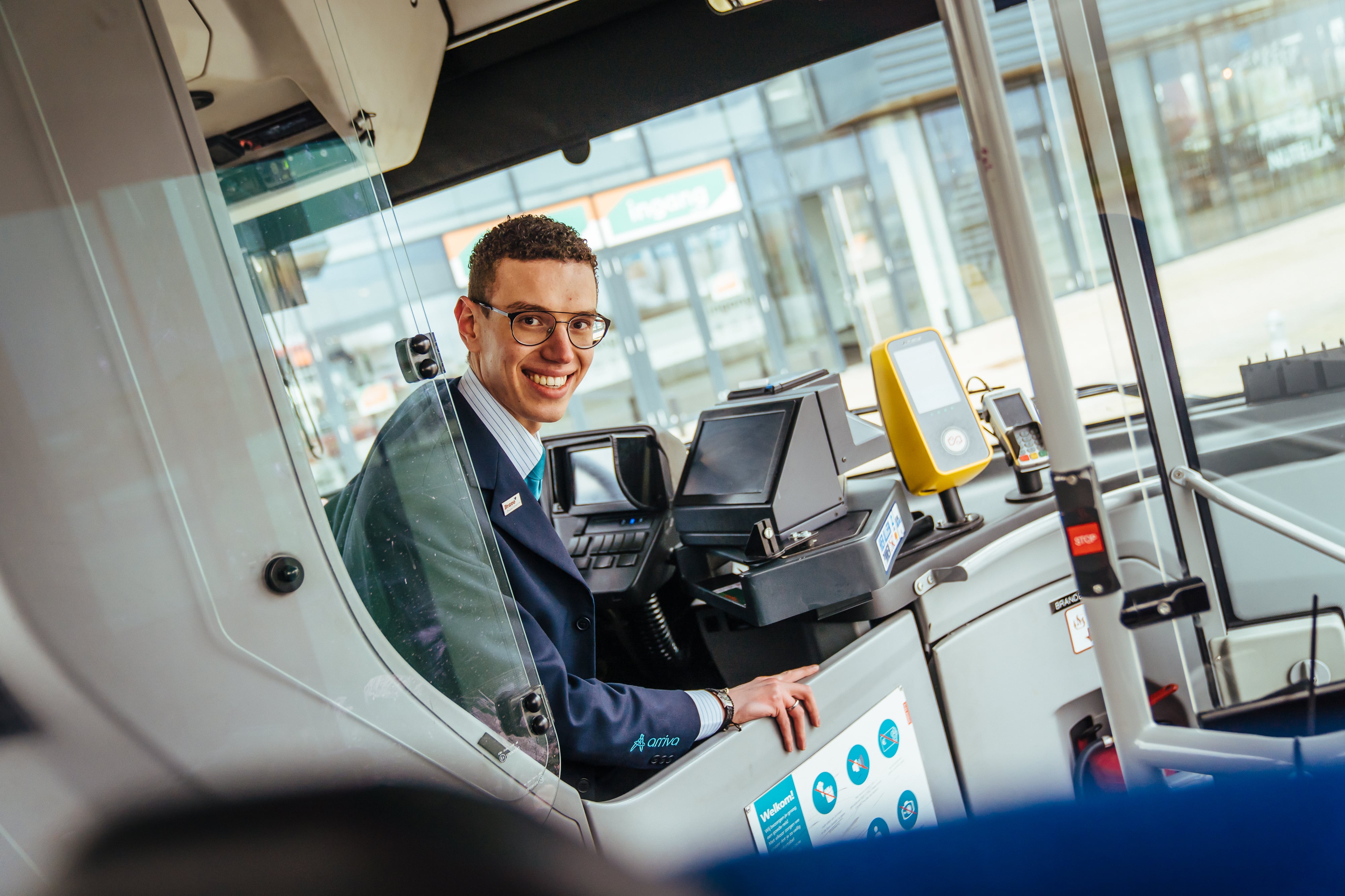 Een mannelijke buschauffeur met een bril zit achter het stuur van een Arriva-bus en kijkt achterom.