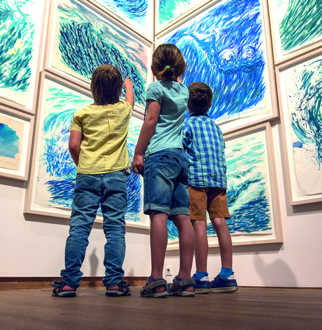 Drie kinderen van een jaar of vijf vanaf de rug gezien staan voor een muur vol met schilderijen in blauw- en groentinten.