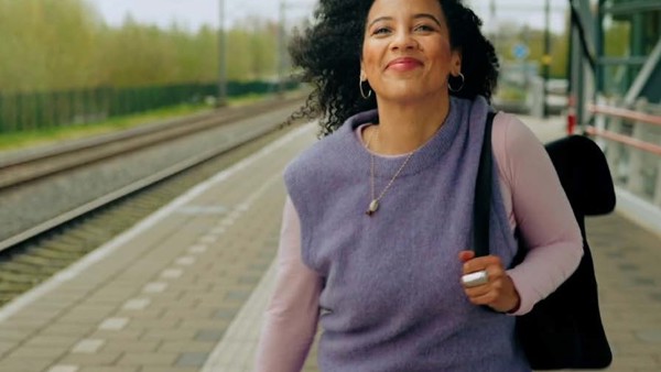 Jonge vrouw staat lachend op een treinperron met een zwarte rugzak duidelijk zichtbaar over haar linkerschouder.