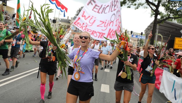 Juichende vrouwelijke deelnemers aan de Vierdaagse zijn net gefinished en dragen hun medaille en hebben gladiolen en een groot spanddoek vast.