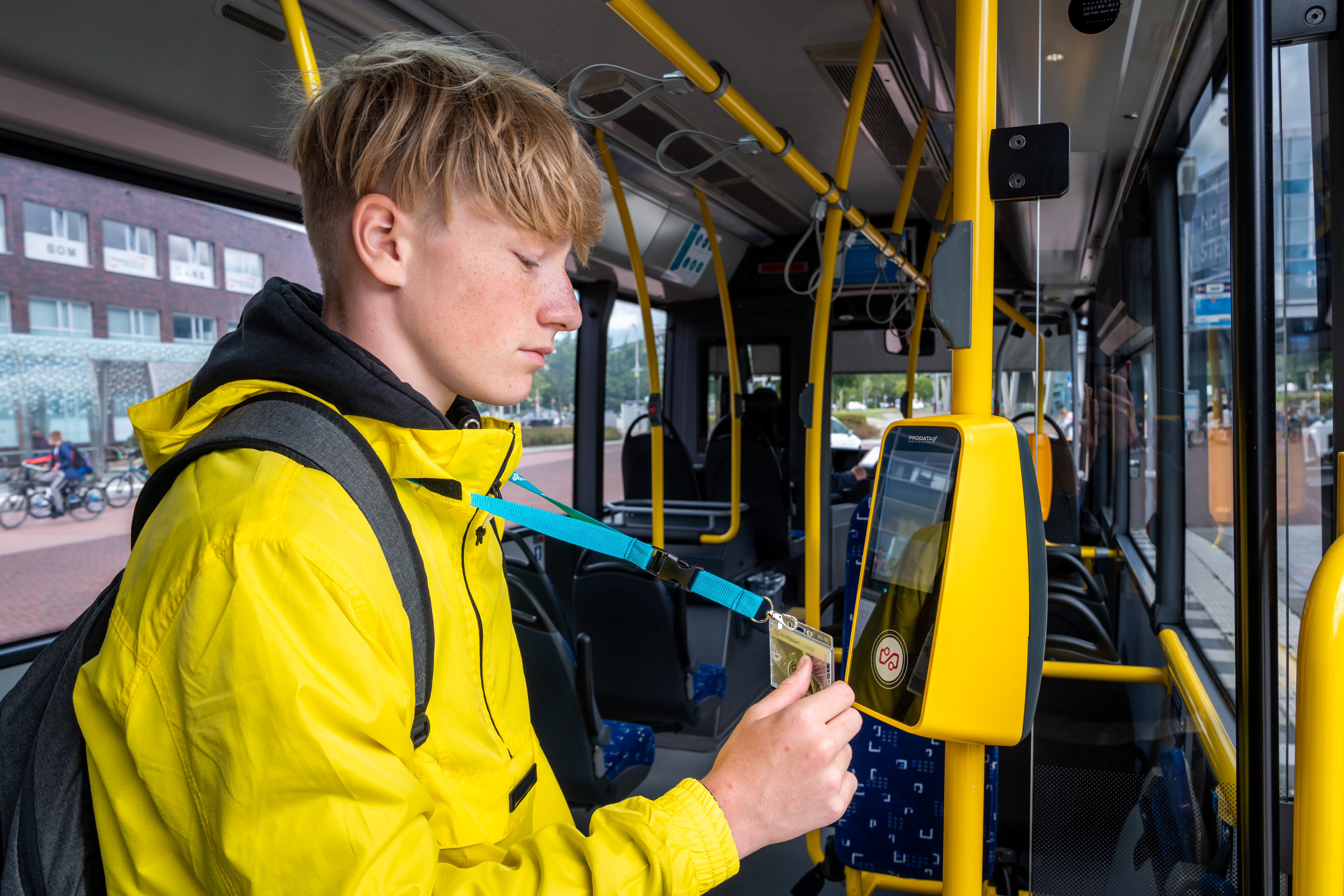 Tiener in een gele jas houdt zijn OV-chipkaart bij de incheckpaal in een bus van Arriva.