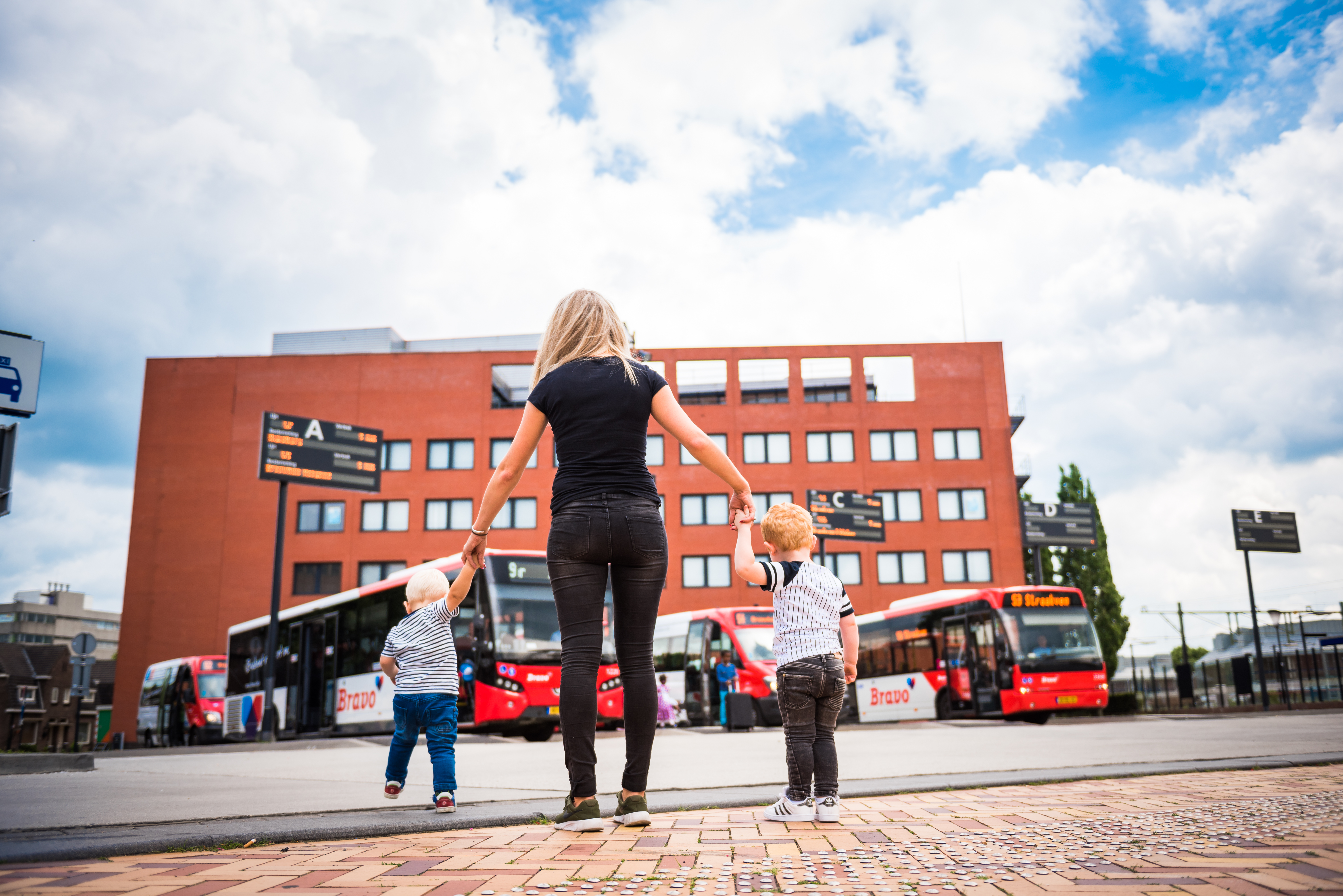 Een jonge vrouw met aan elke hand een klein kindje, vanaf de rug bezien kijk zij uit over een busstation waar rode Arriva-bussen geparkeerd staan.