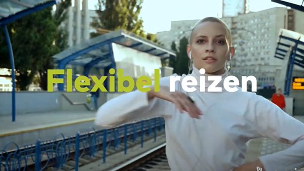 Vrouw met gemillimeterd haar in witte coltrui maakt een dansbeweging en kijkt in de camera terwijl ze op een perron staat. Met de tekst 'Flexibel reizen'.