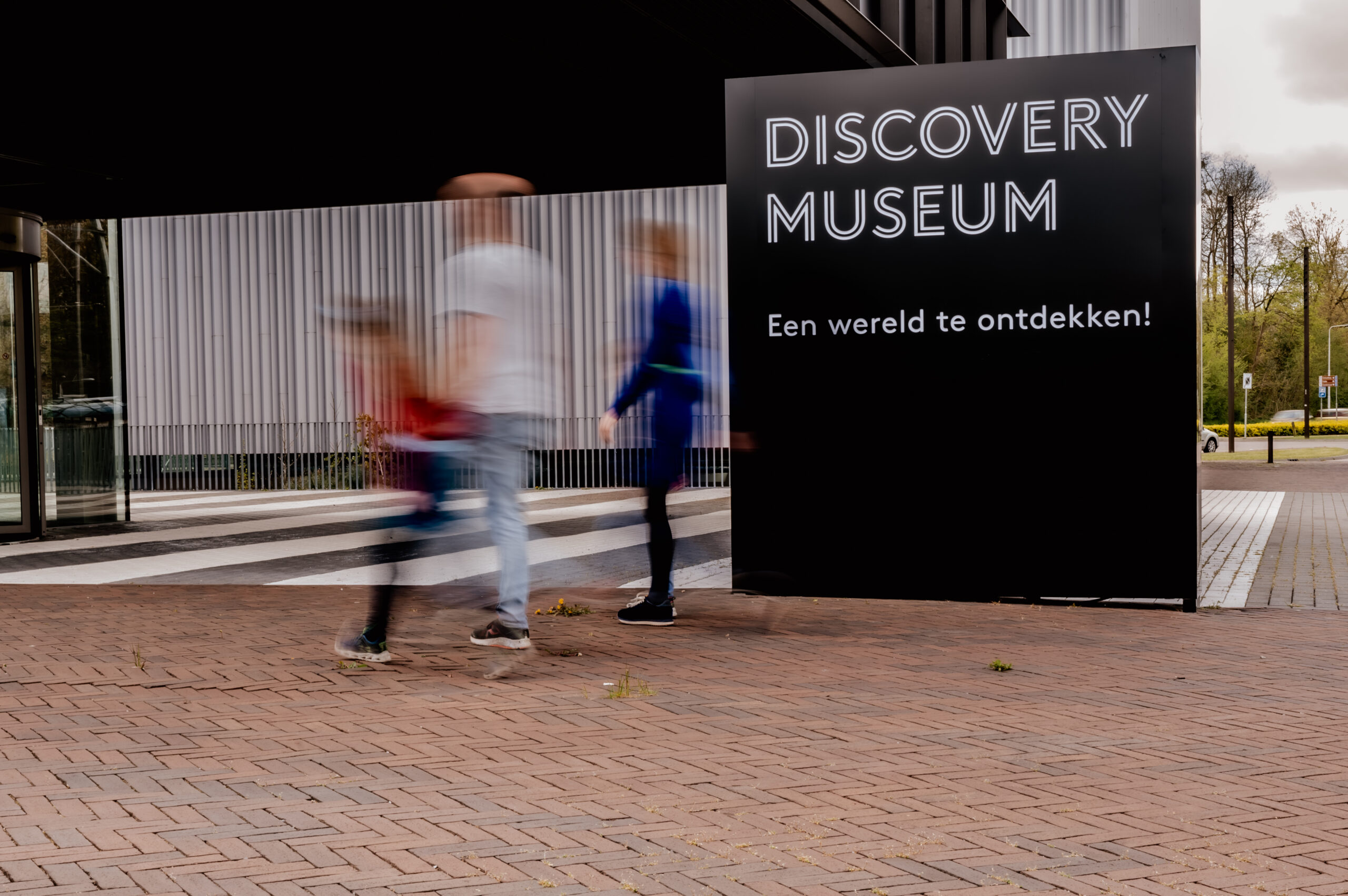 Bordje met daarop 'Discovery Museum, Een wereld te ontdekken!' waar door een lange sluitertijd vervaagd drie mensen voorlangs lopen.
