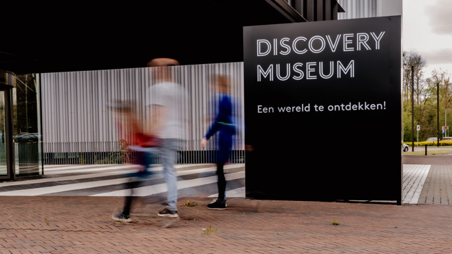 Bordje met daarop 'Discovery Museum, Een wereld te ontdekken!' waar door een lange sluitertijd vervaagd drie mensen voorlangs lopen.