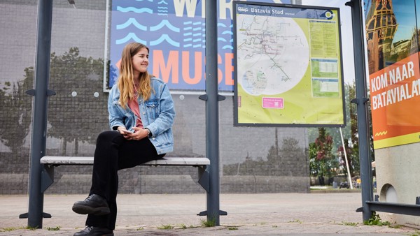 Jonge vrouw zit op bankje van bushalte Batavia Stad met achter haar de vertrekstaat.