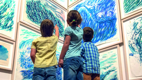 Drie kinderen van een jaar of vijf vanaf de rug gezien staan voor een muur vol met schilderijen in blauw- en groentinten. Drie kinderen van een jaar of vijf vanaf de rug gezien staan voor een muur vol met schilderijen in blauw- en groentinten.