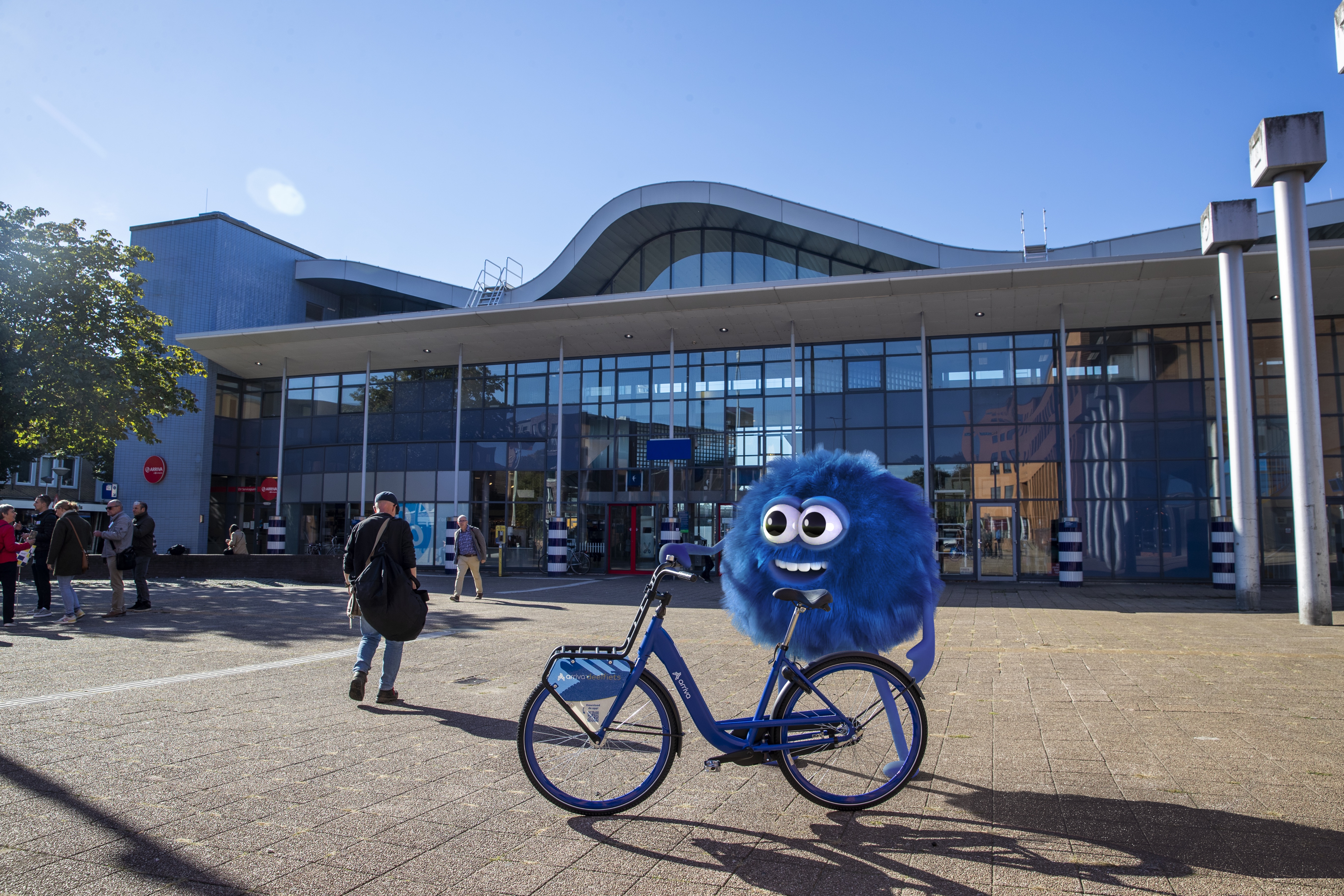 De mascotte van de Arriva-deelfiets, de blauwe fluffy Dirk, staat lachend met zo'n deelfiets voor station Sittard.