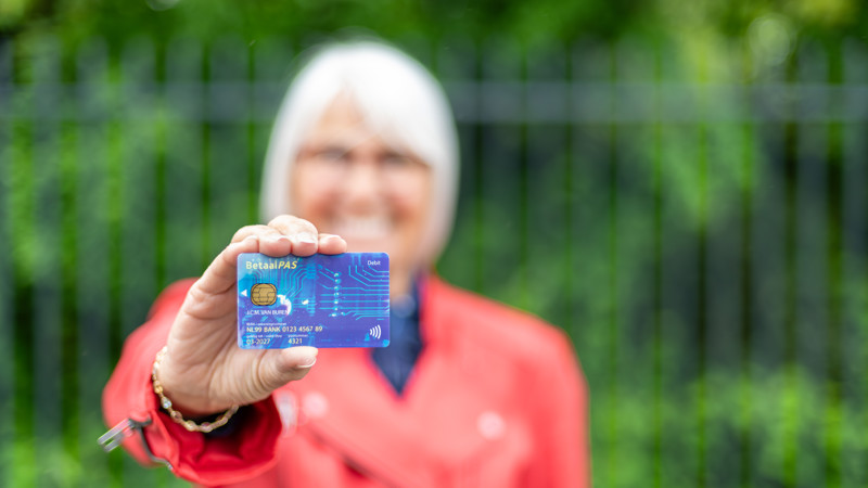 Een oudere vrouw in rode jas houdt een blauwe betaalpas omhoog. Focus ligt op de pas, zij is onscherp op de achtergrond. Een oudere vrouw in rode jas houdt een blauwe betaalpas omhoog. Focus ligt op de pas, zij is onscherp op de achtergrond.