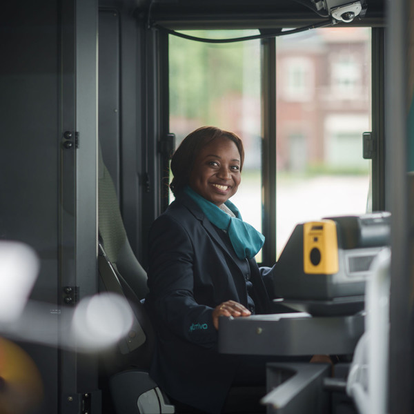 Een glimlachende buschauffeur in Arriva-uniform zit achter het stuur van een bus en kijkt naar de camera. Een glimlachende buschauffeur in Arriva-uniform zit achter het stuur van een bus en kijkt naar de camera.