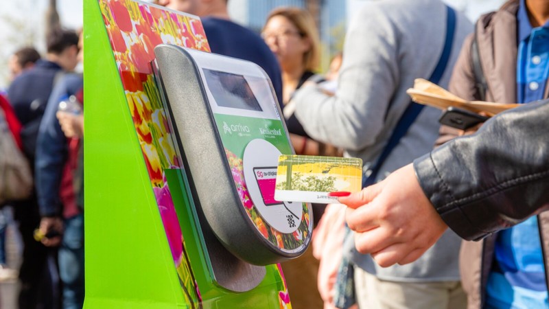 Met kleurige tulpen bestickerde incheckpaal waar een hand een ov-chipkaart bij houdt en met veel mensen in de achtergrond. Met kleurige tulpen bestickerde incheckpaal waar een hand een ov-chipkaart bij houdt en met veel mensen in de achtergrond.