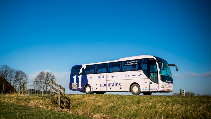 Een blauw-witte touringcar van Arriva rijdt met bestickering van '11 fountains' over een Fries dijkje.