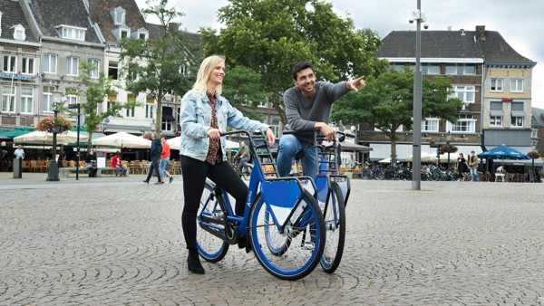 Een man en vrouw staan op een gezellig stadsplein met hun blauwe Arriva-deelfiets, hij wijst en ze lachen allebei.