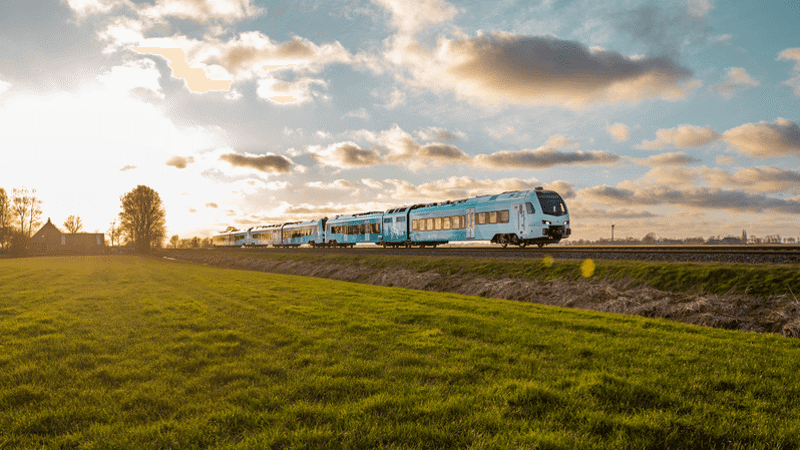 Blauwwitte trein van Arriva rijdt op verhoogd spoor door weilanden in Friesland met boerderij op de achtergrond.