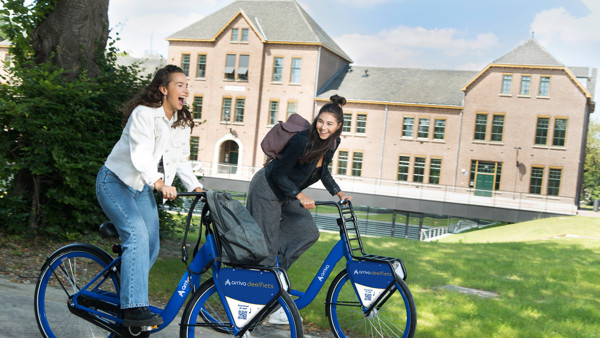 Twee lachende studentes fietsen op blauwe Arriva-deelfietsen door een groene omgeving. Twee lachende studentes fietsen op blauwe Arriva-deelfietsen door een groene omgeving.