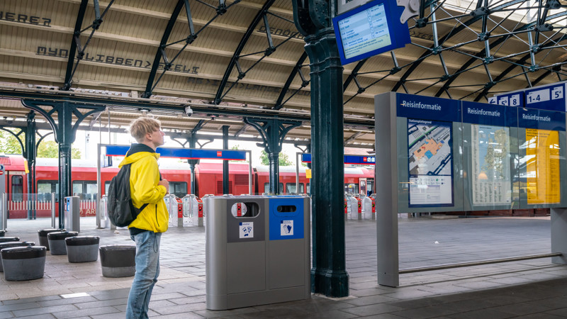 Tiener in felgele jas staat naar de vertrektijden te kijken op een treinperron. Op de achtergrond staat een rode Arriva-trein.