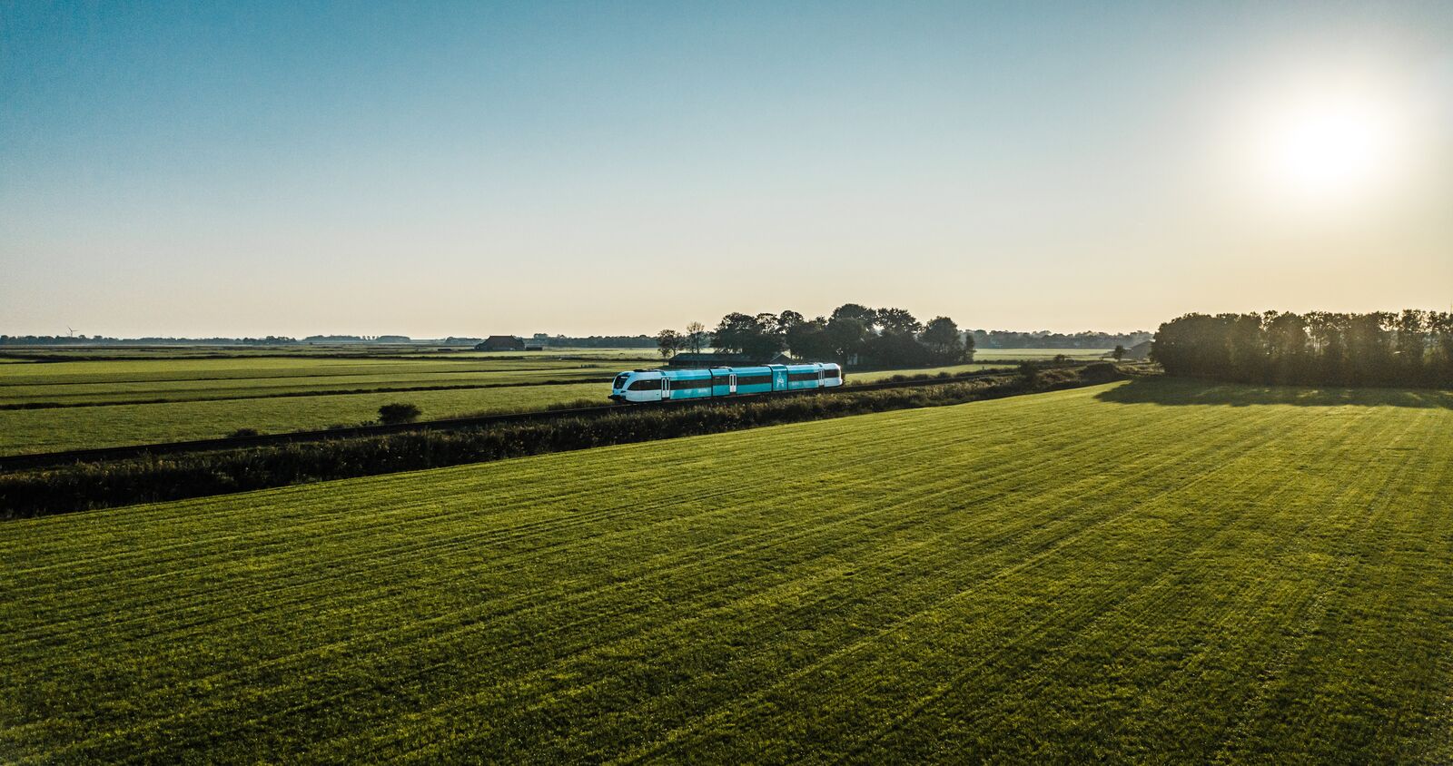 Een Arriva-trein doorkruist een vlak landschap vol weilanden op een stralende dag met strakblauwe lucht.