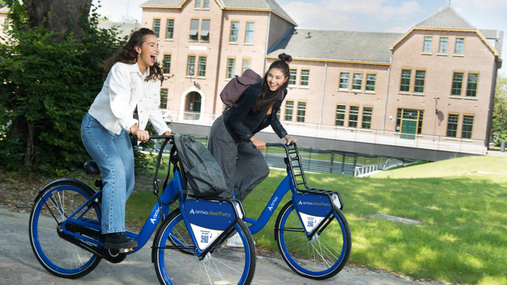Twee jonge vrouwen fietsen lachend op Arriva-deelfiets langs groot huis in groene omgeving.