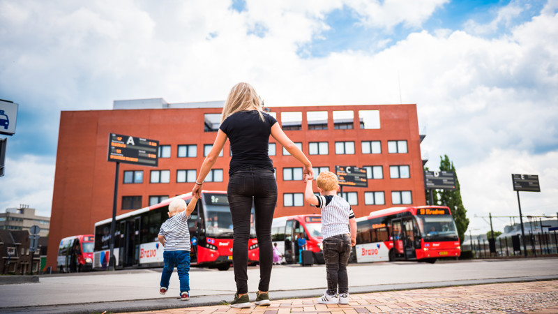 Een jonge vrouw met aan elke hand een klein kindje, vanaf de rug bezien kijk zij uit over een busstation waar rode Arriva-bussen geparkeerd staan.