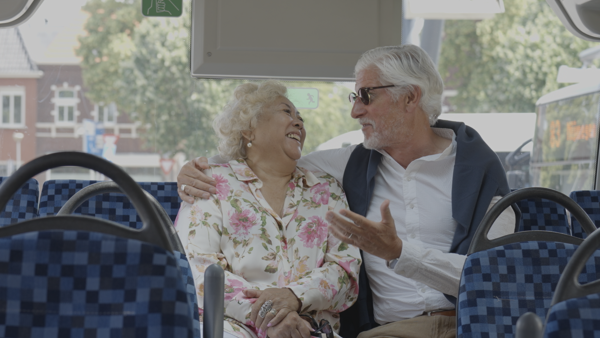 Ouder stel zit achterin Arriva-bus, hij houdt zijn arm om haar schouders, kijkt haar aan en zij lacht breeduit.