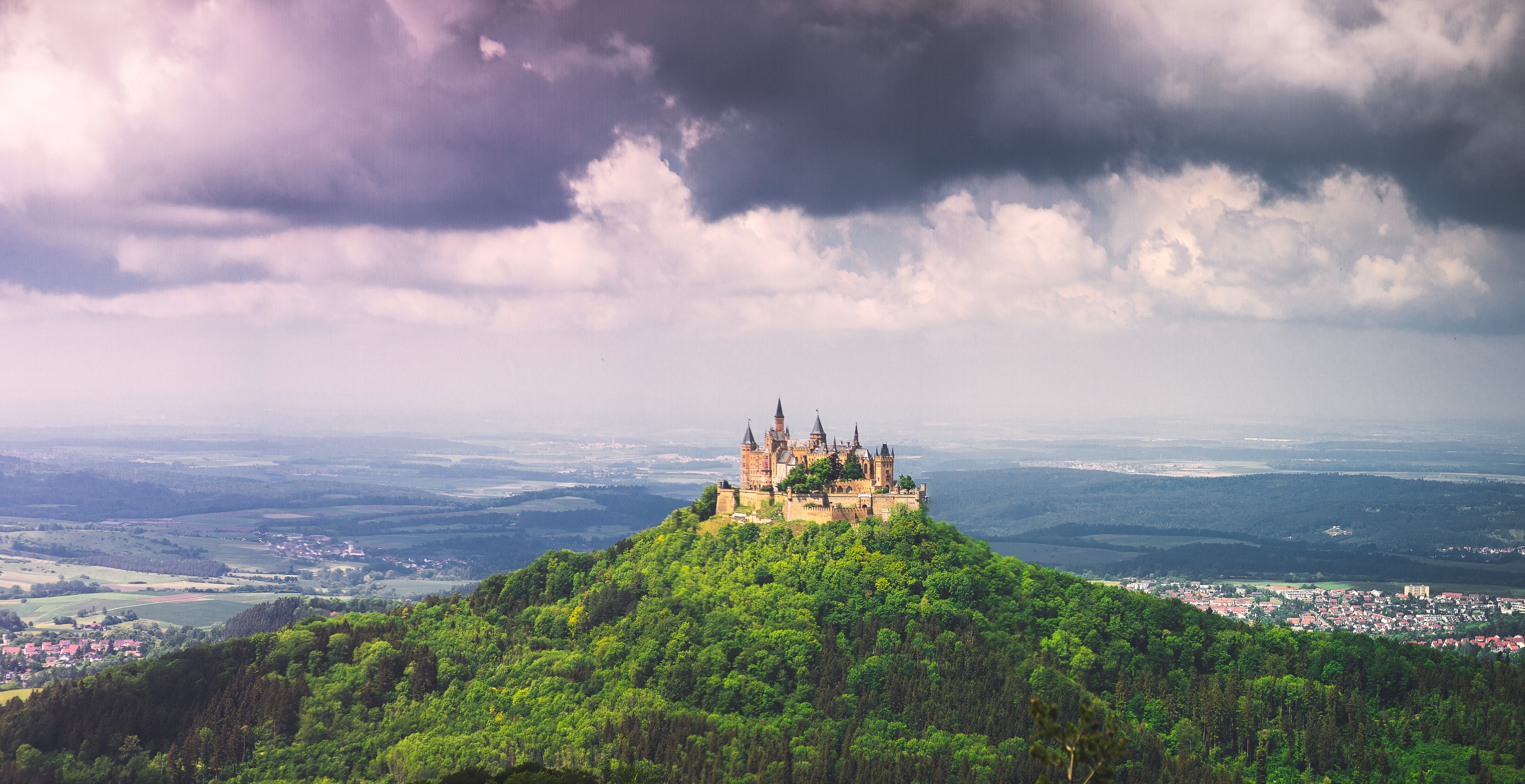 Foto van een kasteel bovenop een groene berg, Hohenzollern, onder een dreigende bewolkte lucht.