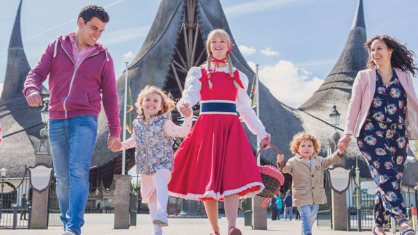 Meisje verkleed als roodkapje loopt met opgetogen jong gezin, hand in hand, de Efteling in, je ziet de ingang achter hen.