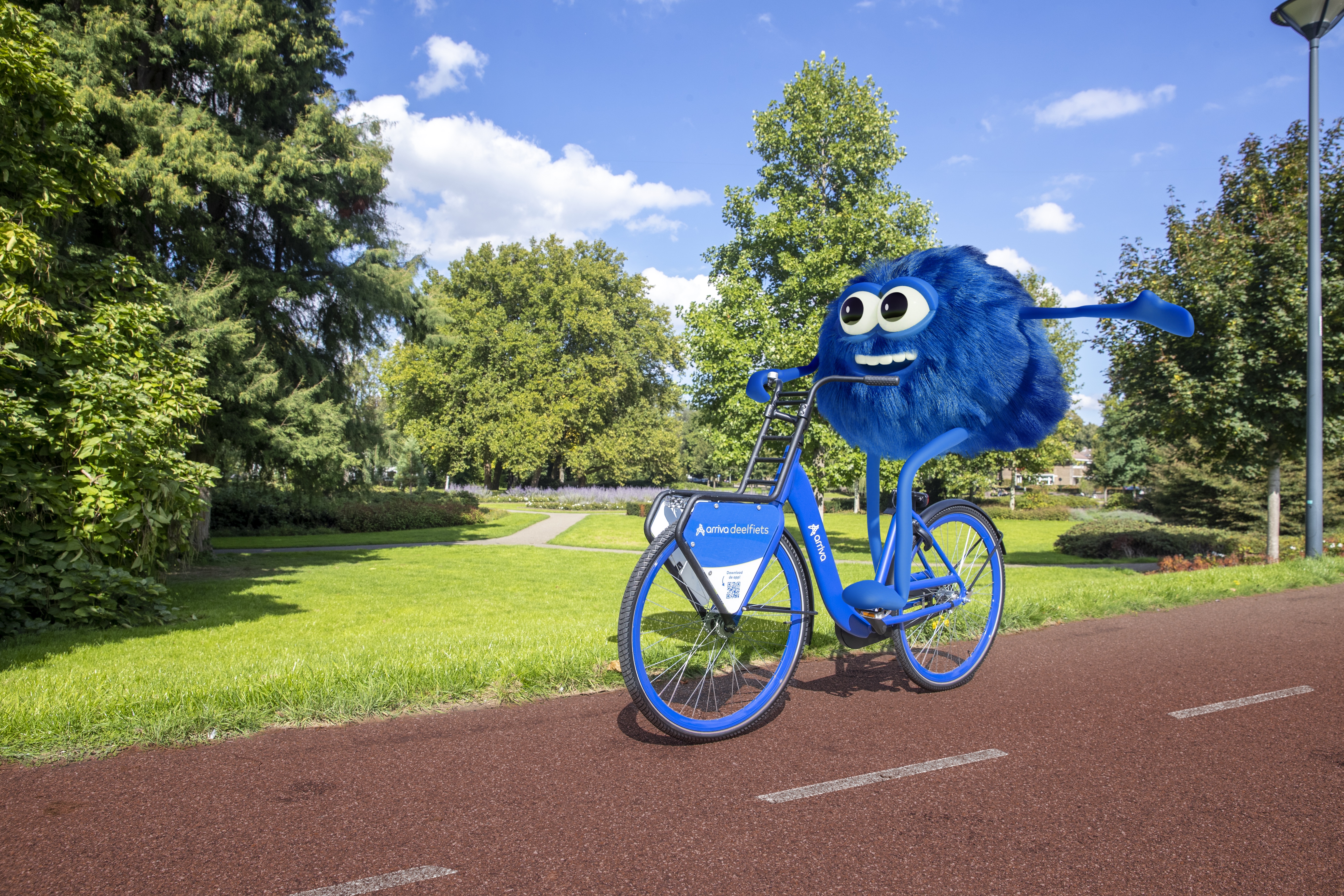 De mascotte van de Arriva-deelfiets, Dirk, zit op zo'n blauwe deelfiets en fiets blij door een stadspark.