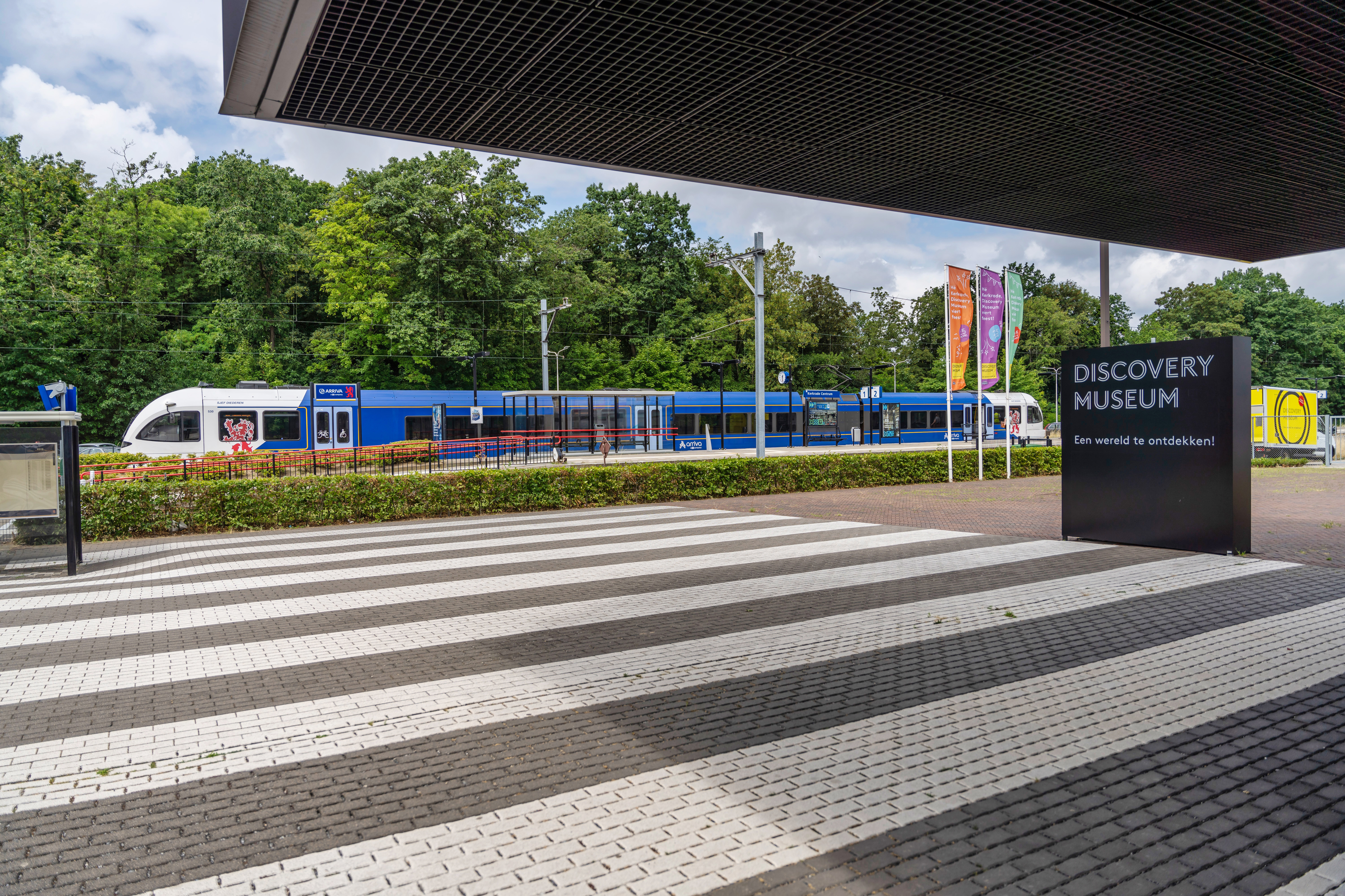 Blauwwitte trein van Arriva staat aan perron van station Kerkrade, op de voorgrond een zebrapad en een bordje met 'Discovery Museum'.