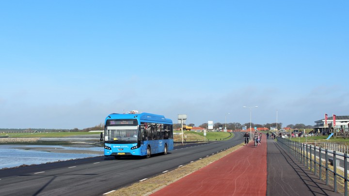 Een blauwe Arriva-bus rijdt over de dijk op Ameland onder een strakblauw hemel naast een fietspad met fietsers.