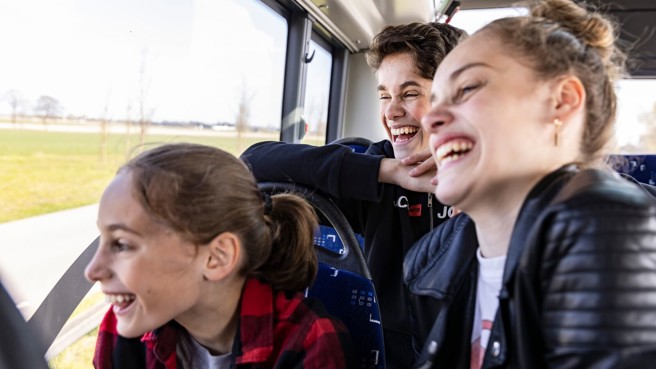 Drie tieners, twee meisjes en een jongen, zitten in een bus van Arriva en moeten om iets lachen dat ze buiten zien.