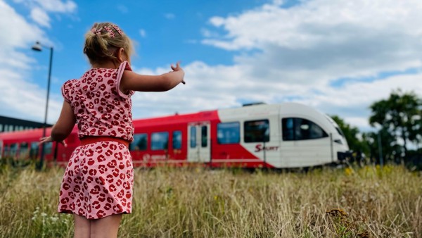 Een meisje van een jaar of vijf in roze zomerjurk staat in hoog gras en kijkt naar een rode Arriva-trein.