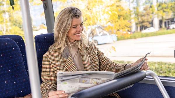 Jonge vrouw in geruit jasje zit lachend in de bus de krant te lezen.