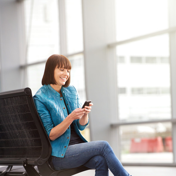 Een jonge vrouw houdt haar telefoon vast en lacht, ze zit met haar benen over elkaar op een bankje in een wachtruimte Een jonge vrouw houdt haar telefoon vast en lacht, ze zit met haar benen over elkaar op een bankje in een wachtruimte