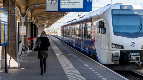 Reiziger loopt over perron van een station langs een blauwwitte trein van Arriva die naar Roermond gaat.