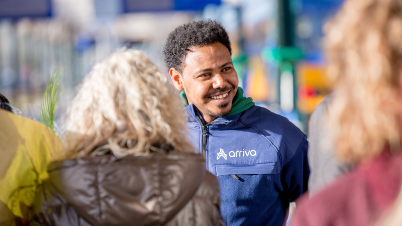 Lachende man in Arriva-jas, een medewerker van het Serviceteam, omringd door mensen, waaronder een vrouw met bloemen. Lachende man in Arriva-jas, een medewerker van het Serviceteam, omringd door mensen, waaronder een vrouw met bloemen.