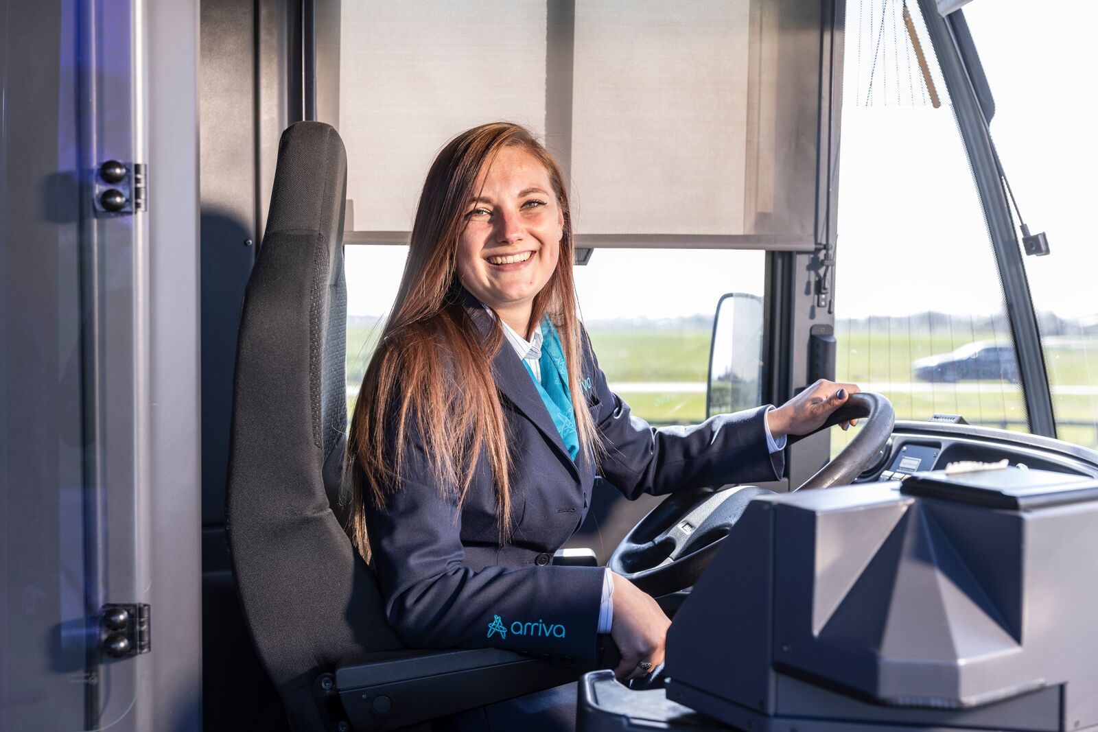 Jonge, vrouwelijke buschauffeur in uniform zit lachend achter het stuur van Arriva-bus.