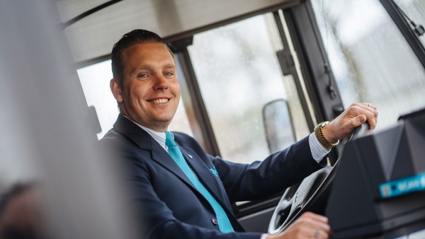 Buschauffeur in uniform zit achter het stuur van zijn Arriva-bus en kijkt lachend naar buiten.